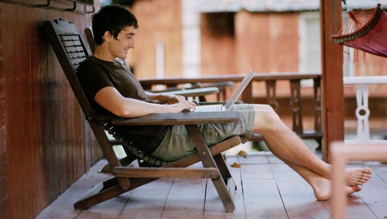 Man sitter i solstol utanför en bungalow och jobbar på sin dator