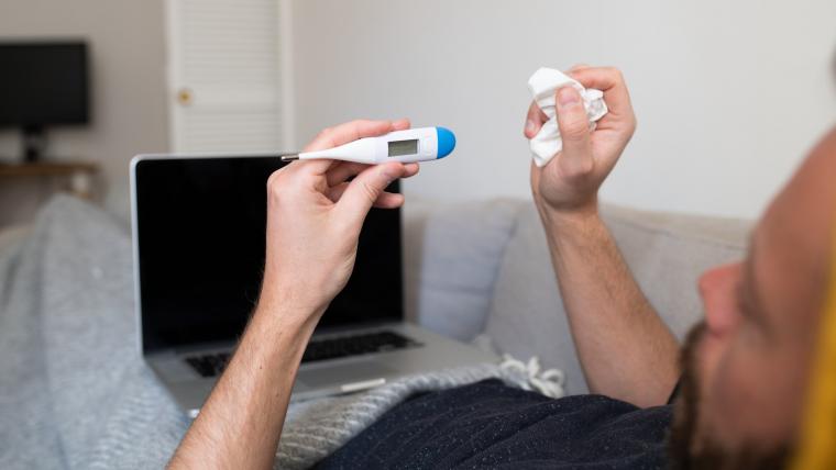 Man ligger i säng med dator på magen och med en febertermometer i handen